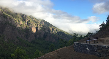 Blick aus Richtung Eingang zum Parque Nacional de la Caldera de Taburiente in Richtung Cumbre Nueva, ein etwa 10 km langer Höhenzug, der den Ostteil vom Westteil der Insel La Palma trennt. Ein typisches Phänomen wird als Cascada de nubes („Wolkenwasserfall“) bezeichnet. In einer Höhe von etwa 1450 Metern drängen sich aus Osten kommende Passatwolken über den Bergkamm und fallen auf der Westseite herunter, wobei diese sich dann auflösen.